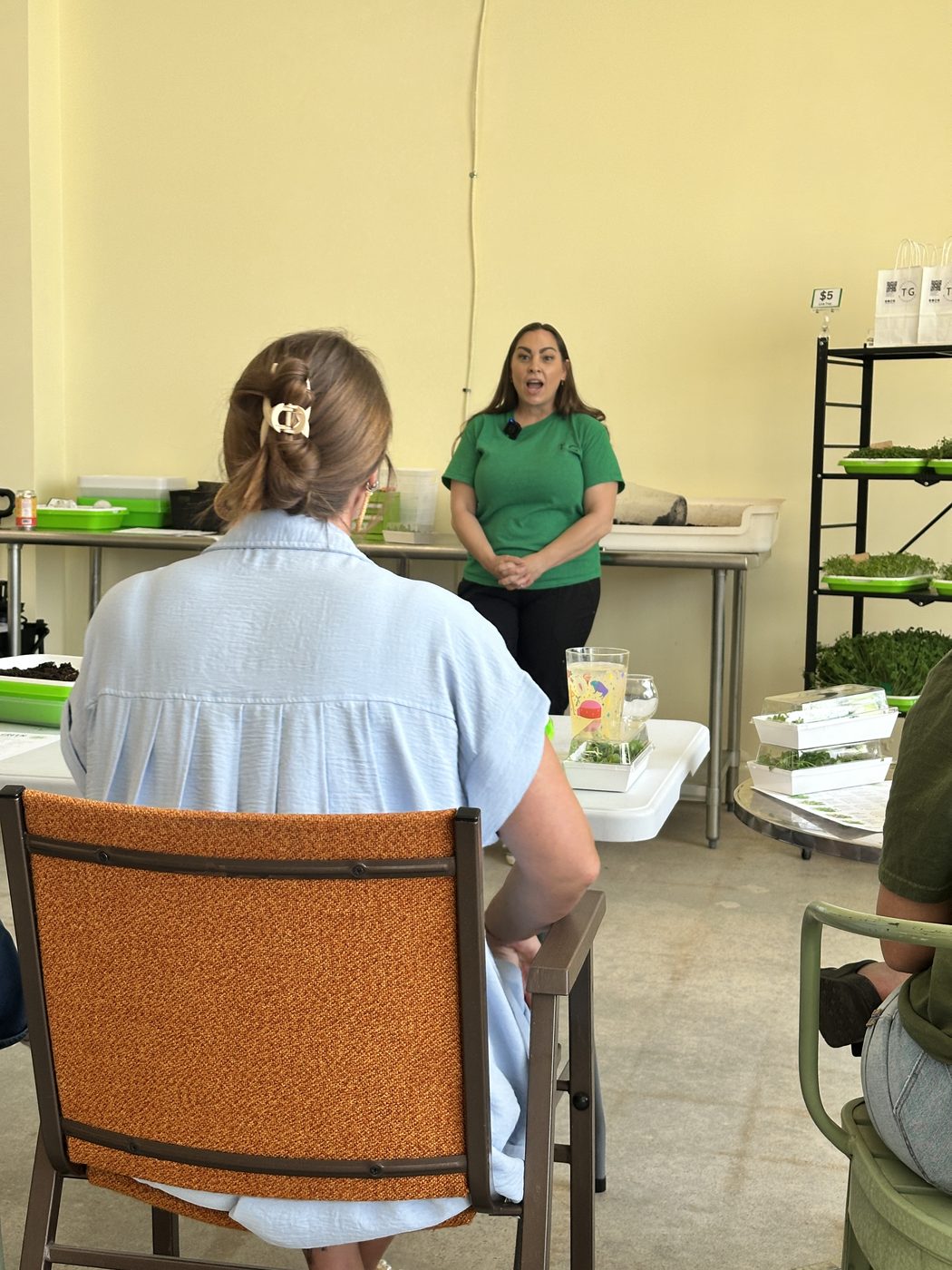 Brooke talking through microgreen growing to a seated attendee, trays of greens on the rack behind her